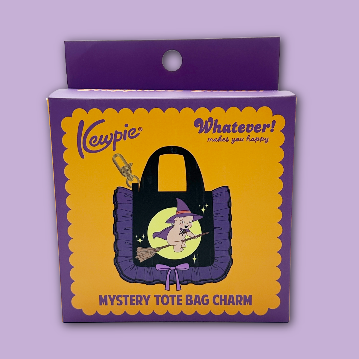 Kewpie 'Whatever! Makes you happy' mystery tote bag charm showing a witch-themed tote bag in a purple and yellow cardboard package, displayed against a light purple background.