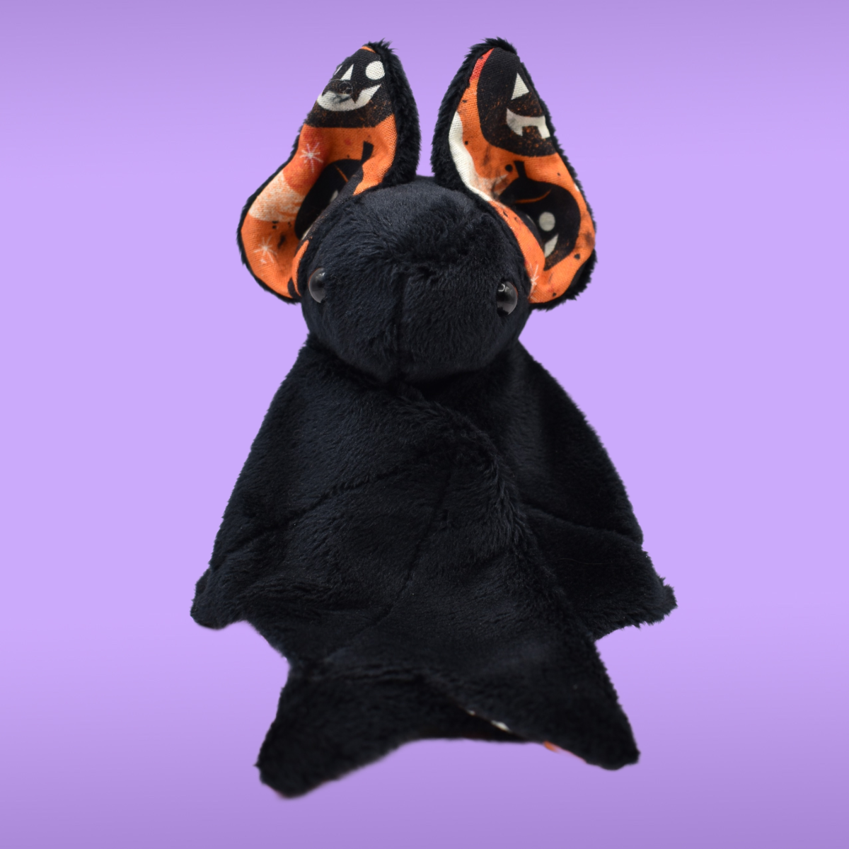 Black Spookling bat plush with orange Halloween print ears against a lavender background, shown with folded wings