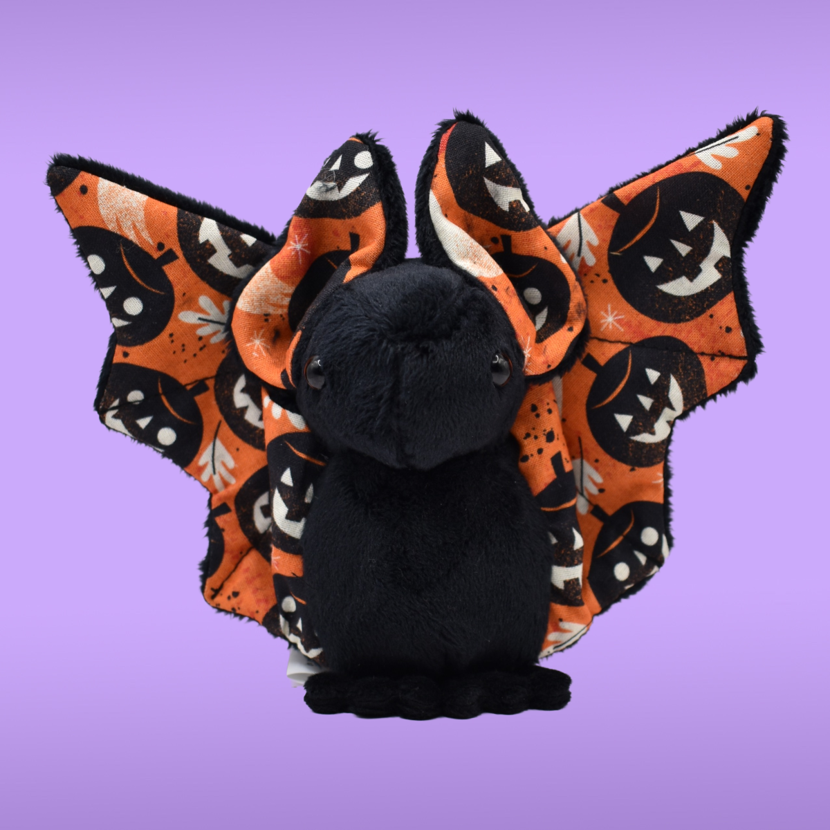 Black Spookling bat plush with fully open orange Halloween print wings displayed on a lavender background