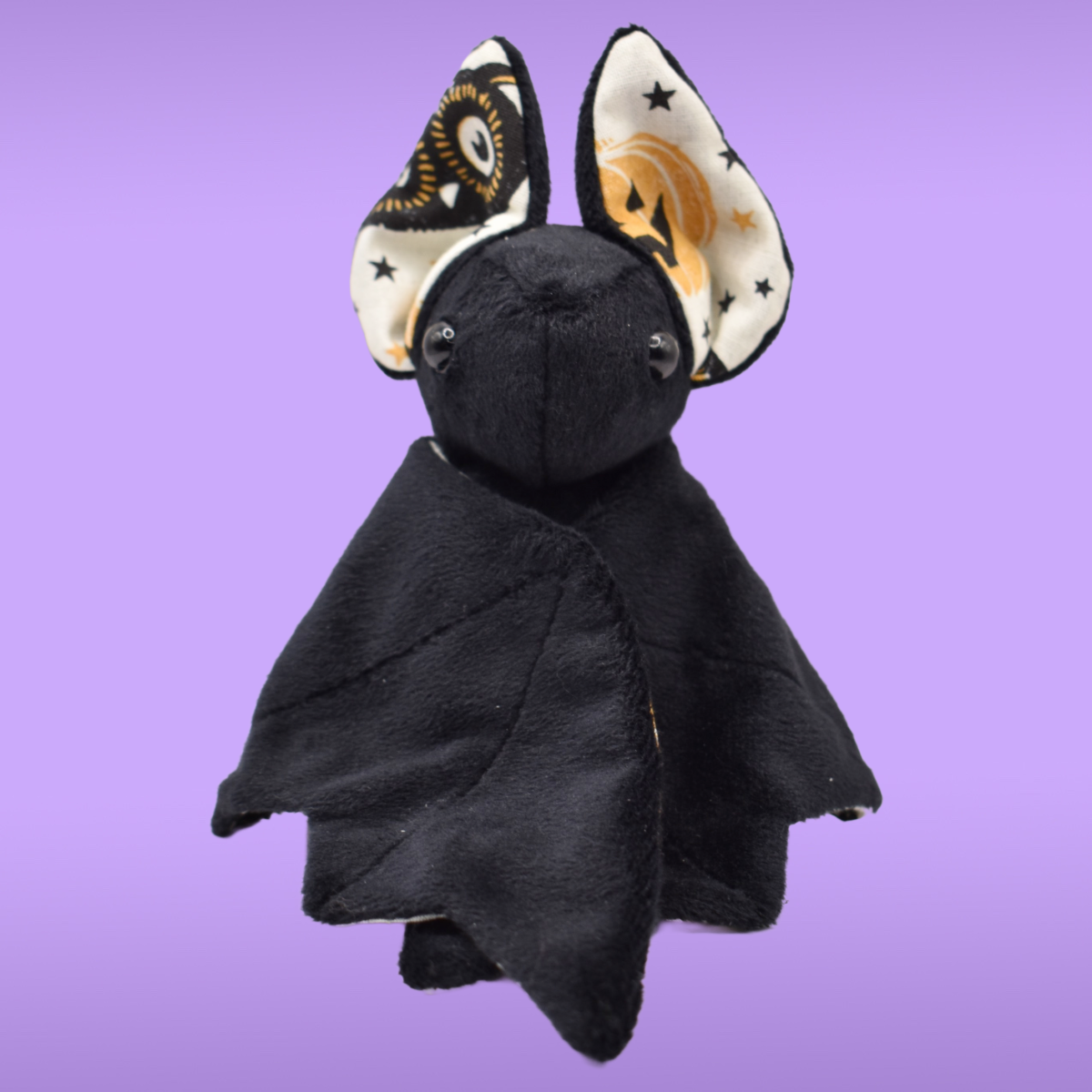 Black Spookling bat plush with vintage Halloween print ears against a lavender background, shown with folded wings.