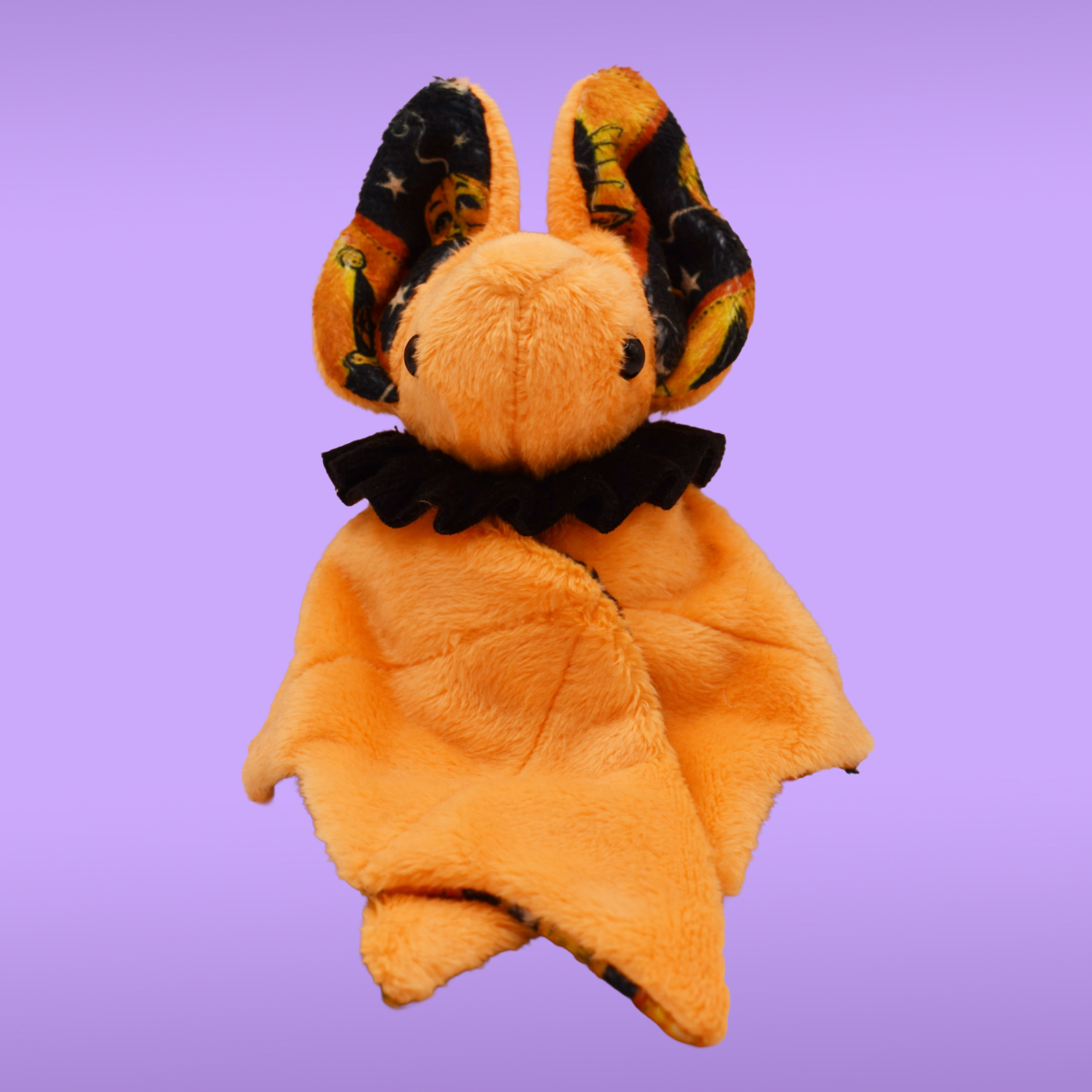 Orange Spookling bat plush with black ruffle collar and folded wings against a lavender background.