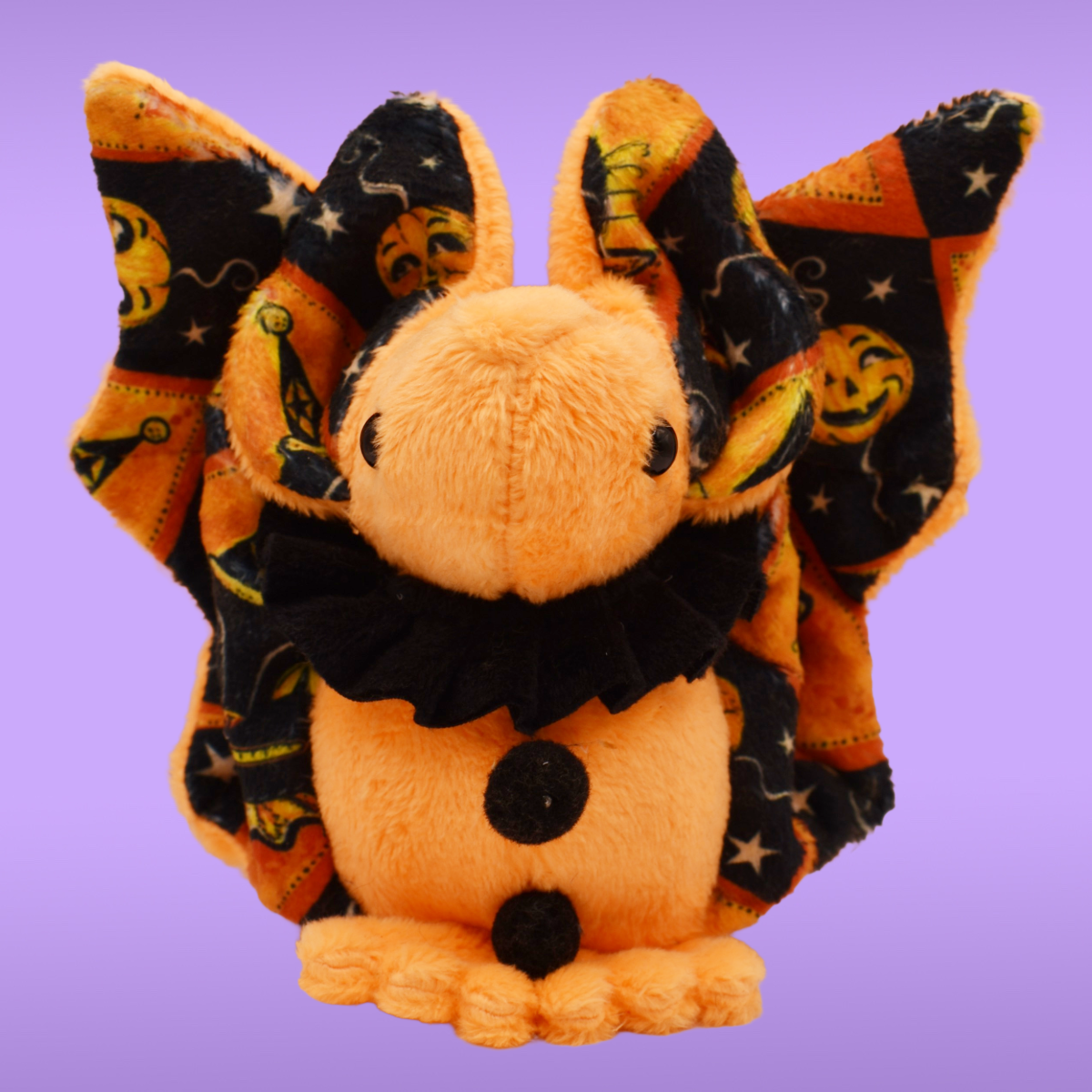 Orange Spookling bat plush with vintage Halloween print wings, black ruffle collar, and open wings displayed against a lavender background.